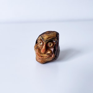 Könnte beinhalten: Ein kleines, geschnitztes Holzgesicht mit detailliertem, ausdrucksstarkem Design. Das Gesicht hat eine markante Nase, große Augen und einen lächelnden Mund. Die Farben sind hauptsächlich Braun und Beige, mit dunkleren Akzenten. Das Objekt scheint ein Dekorationsartikel oder eine Figur zu sein.