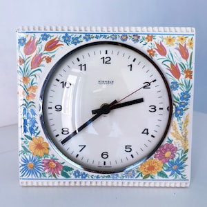 May include: Square ceramic clock with a white face and black hands. The clock face has black numbers and the brand name "Kienzle Automatic". The frame is decorated with colorful flowers and foliage.
