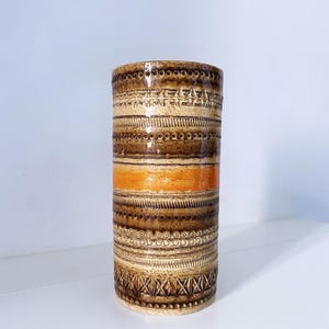 Op de afbeelding: Een cilindrische keramische vaas met een getextureerd oppervlak met horizontale banden van bruin en beige glazuur. Een opvallende oranje streep omcirkelt het midden, terwijl de buitenkant is versierd met herhalende geometrische patronen, waaronder stippen, lijnen en kruisarceringen.