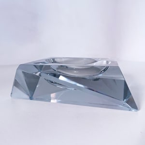 Op de afbeelding: Een glazen asbak met een geometrisch ontwerp. De asbak is piramidevormig met een vierkante basis en een afgeronde bovenkant.