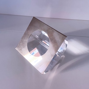 May include: A silver metal square with a round hole in the center, sitting on a clear acrylic base. The base is angled and has a reflective surface.