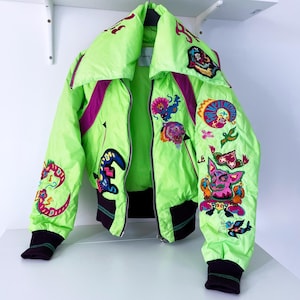 Puede incluir: Una chaqueta bomber verde brillante con ribetes rosa y morado. La chaqueta está decorada con diseños bordados coloridos, incluyendo flores, animales y patrones geométricos.
