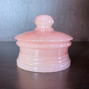 Puede incluir: Una caja de alhajas de alabastro rosa con una tapa redondeada y una banda decorativa alrededor de la base.