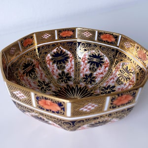 Può includere: Una ciotola ottagonale in ceramica con bordo dorato e intricati motivi floreali. La ciotola presenta uno sfondo blu navy con disegni floreali arancioni e bianchi. Il bordo dorato aggiunge un tocco di eleganza.