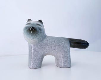 May include: A ceramic cat figurine with a speckled gray body and a dark brown tail. The cat has a rounded head with small ears and a slightly upturned nose. The figurine is a minimalist design.