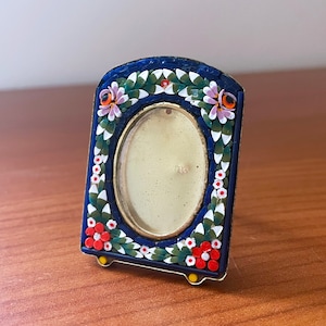 May include: A small, blue and gold picture frame with a floral mosaic design. The frame has a white oval opening for a photo.