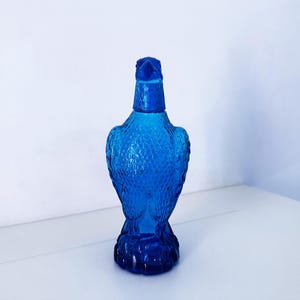 Könnte beinhalten: Eine kobaltblaue Glasflasche in Form eines Adlers. Die Flasche hat eine strukturierte Oberfläche und ein detailliertes Design, einschließlich des Kopfes, der Flügel und der Füße des Adlers. Sie steht auf einer weißen Oberfläche. Die Flasche ist etwa 25 cm hoch.
