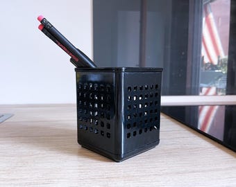 May include: A black, square pencil holder with a grid of square cutouts. Two black pencils with pink erasers are inside. The holder sits on a light wood surface, with a blurred background.
