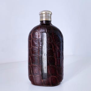 Peut inclure: Une flasque en cuir marron avec un dessus argenté et un insert en métal. La flasque a un motif en peau de crocodile.