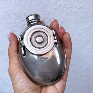 Puede incluir: Una pequeña petaca ovalada de plata con una tapa circular con bisagras. La petaca tiene un tapón de rosca y un elemento circular decorativo en la parte delantera. El texto "REV: 1535" está estampado en la petaca. La petaca se sostiene en una mano.