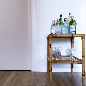Puede incluir: Un carrito de bar de bambú de dos niveles con botellas de licor y vasos. La estantería superior contiene varias botellas de ginebra, vodka y otros licores. La estantería inferior tiene copas de cóctel. El carrito es de bambú marrón claro, de unos 60 cm de altura.