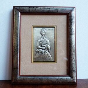 May include: A silver framed picture with a silver relief sculpture of a woman in a traditional dress. The sculpture is set against a beige background and the frame has a wood-like finish.