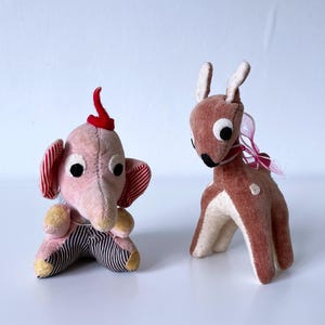 Può includere: Due giocattoli di peluche vintage: un elefante rosa con orecchie a righe rosse e bianche e un cappello rosso, e un cervo marrone con un nastro rosa. L'elefante indossa pantaloni a righe. Entrambi i giocattoli hanno occhi neri.