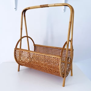 May include: A vintage rattan magazine rack with a handle. The rack is made of woven rattan and has a rectangular shape with rounded ends. The rack is approximately 18 inches long, 12 inches wide, and 15 inches tall.