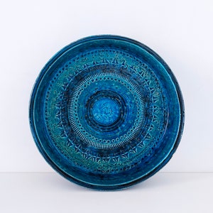 May include: A circular ceramic plate with a textured surface and a deep blue glaze. The plate features concentric circles with detailed patterns, including geometric shapes and dotted lines. The centre has a raised, floral-like design.