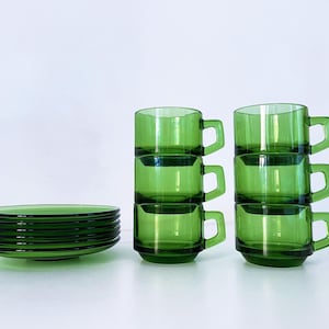Pode incluir: Um conjunto de chávenas e pires de vidro verde vintage. As chávenas são empilhadas em duas colunas de três, e os pires são empilhados separadamente. A loiça tem um design simples e geométrico e uma cor verde vibrante.