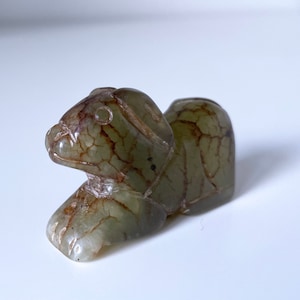 May include: A small, green jade sculpture of a dog lying down. The sculpture has intricate details and a mottled green and brown color.