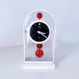 May include: A white and clear retro-style clock with a black clock face and white hands. The clock has red accents on the top and bottom. The clock is sitting on a white surface.