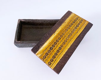 May include: A rectangular ceramic box with a lid. The box is brown with a gold geometric pattern on the lid.