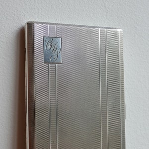 May include: Silver cigarette case with a monogrammed initials "C.D." on the front. The case has a textured, striped design.
