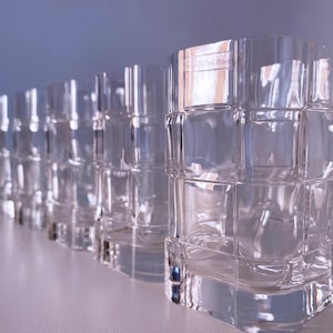 May include: A row of clear, square-patterned drinking glasses. The glasses are made of transparent glass and are arranged in a line, with the closest glass in focus. The glasses have a textured, grid-like design.