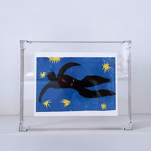 Könnte beinhalten: Ein gerahmter Druck einer schwarzen Silhouette einer Person, die auf dem Rücken auf einem blauen Hintergrund mit gelben Sternen liegt.