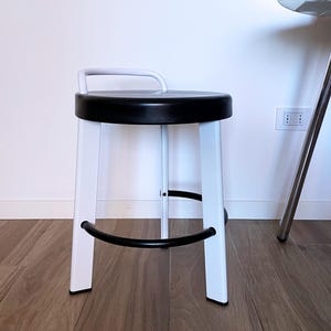 Könnte beinhalten: Ein weißer Metallhocker mit einem schwarzen gepolsterten Sitz und einer schwarzen Fußstütze aus Metall. Der Hocker hat eine gebogene Rückenlehne.