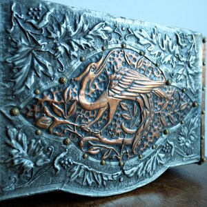 May include: A decorative metal wall plaque featuring a copper bird design. The bird is surrounded by leaves and vines, and the plaque is accented with copper rivets.