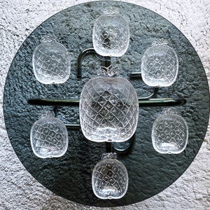 May include: A set of seven clear glass dishes shaped like pineapples, consisting of one large central dish and six smaller surrounding dishes, all featuring a textured, cross-hatched surface pattern that mimics the skin of a pineapple.