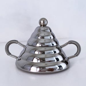 May include: A silver sugar bowl with a lid and two handles. The bowl has a beehive-like design with a small ball on top of the lid. The handles are a coiled design.