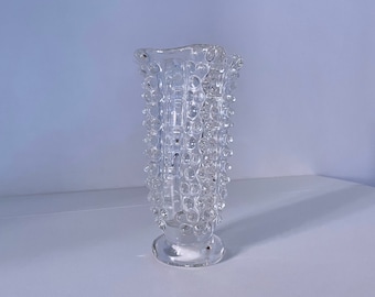 May include: Clear glass vase with a textured, bubble-like design. The vase has a flared top and a round base. The clear glass allows light to pass through, highlighting the unique texture. A decorative piece for home decor.
