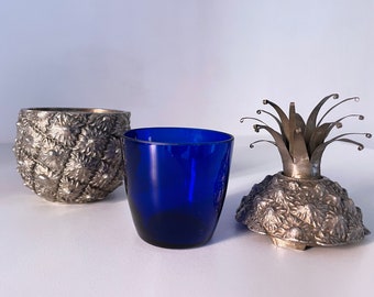 May include: A silver pineapple shaped container with a blue glass lid. The container is decorated with a textured pineapple design. The lid has a metal pineapple top.