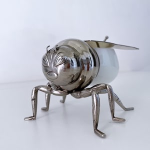 Op de afbeelding: Decoratief metalen bijenbeeldje met een glanzende zilveren afwerking. De bij heeft een afgerond lichaam, zes poten en een paar vleugels. Het lichaam van de bij bestaat uit een zilveren kop en een wit, eivormig lichaam. Een uniek woondecoratie-item.