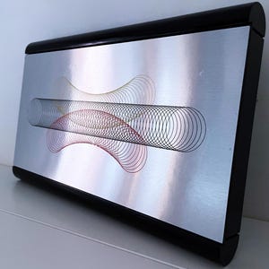 May include: A silver metal wall art piece with a black frame. The art features a colourful, abstract design of overlapping circles and lines.