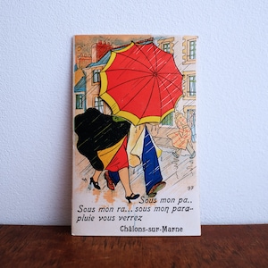 Op de afbeelding: Een vintage ansichtkaart met een cartoonillustratie van twee mensen die in de regen lopen. De persoon vooraan houdt een rode paraplu met een gele rand vast. De tekst op de ansichtkaart luidt "Sous mon pa... Sous mon ra... sous mon para-pluie vous verrez Châlons-sur-Marne".