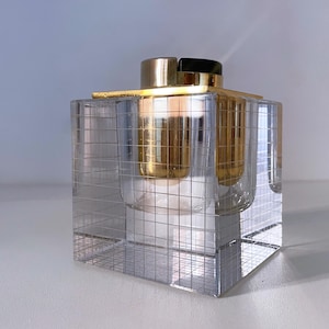 May include: A clear glass cube with a grid pattern, containing a gold metal lighter with a black top.