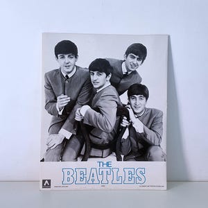 May include: A black and white photograph of The Beatles, a British rock and pop band. The band members are wearing matching suits and have short, dark hair. The band's name is printed in blue letters at the bottom of the image.