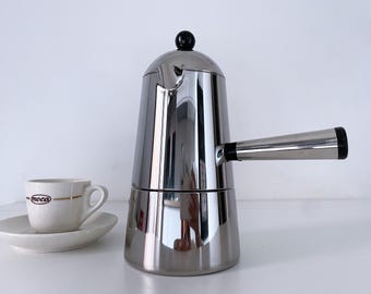May include: A stainless steel stovetop espresso maker with a black handle and knob, next to a small white cup and saucer. The cup has the word "moca" printed on it.