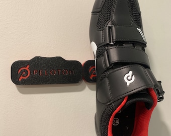 May include: Black cycling shoes with three velcro straps and a red interior. The shoes have a white logo on the side and a black and red Peloton logo on the bottom.