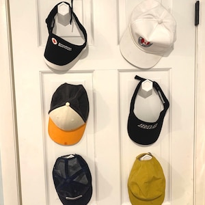 May include: Six hats hanging on hooks on a white door. The hats include a black and white American Red Cross hat, a white hat with a red logo, a black and white hat with an orange brim, a black hat with a white brim, a blue and black mesh hat, and a yellow hat.