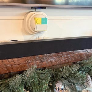 May include: A black soundbar rests on a rustic wooden mantel decorated with evergreen garland and ornaments. Above, a television is mounted on the wall. A small, wall-mounted device with yellow and teal accents is visible.