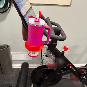 Könnte beinhalten: Ein pinkfarbener Stanley Quencher H2.0 FlowState-Becher mit weißem Deckel und Griff ist an einer roten Halterung befestigt, die an einem schwarzen Peloton-Heimtrainer angebracht ist. Der Becher ist mit "QUENCHER H2.0 TUMBLER" beschriftet.