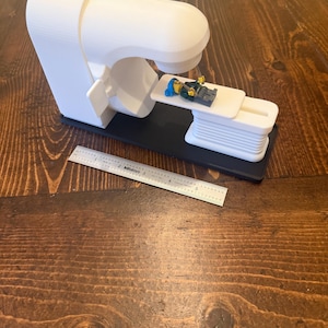 Peut inclure: Modèle 3D imprimé en blanc d'une machine d'IRM avec une figurine Lego allongée sur la table à l'intérieur. Le modèle est sur une base noire et une règle se trouve au premier plan.