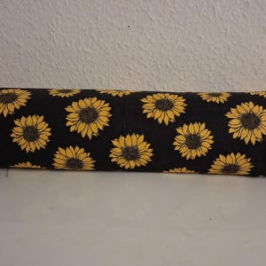 May include: A rolled-up fabric featuring a repeating pattern of yellow sunflowers with black centers on a black background. The sunflowers are in full bloom, with detailed petals. The fabric is likely used for sewing or crafting projects.