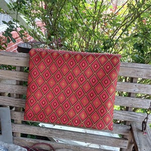 May include: A red and gold patterned cushion with a diamond design sits on a wooden porch swing.
