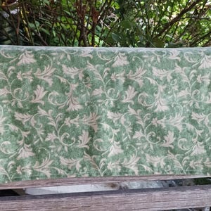 May include: A rectangular, green fabric with a beige floral pattern. The fabric is draped over a wooden bench. The floral design features swirling leaves and stems, creating a classic, elegant look. The fabric's texture appears soft and slightly textured.