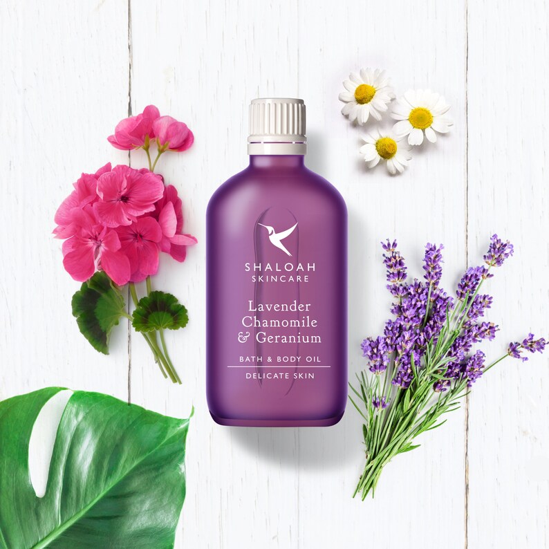 Peut inclure: Bouteille violette d'huile de bain et de corps avec le texte "Shaloah Skincare Lavender Chamomile & Geranium Bath & Body Oil Delicate Skin". La bouteille est entour&eacute;e de fleurs de lavande, de camomille et de g&eacute;ranium.