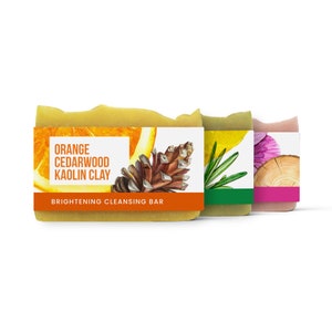 Peut inclure: Trois savons avec des parfums différents. Le premier savon est jaune et porte une étiquette indiquant "Orange Cedarwood Kaolin Clay Brightening Cleansing Bar". Le deuxième savon est vert et porte une étiquette avec un brin de romarin. Le troisième savon est rose et porte une étiquette avec une fleur.