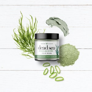 May include: A green jar of Dead Sea Minerals Clay Mask with a black lid. The jar is surrounded by green seaweed, green powder, and green aloe vera slices on a white wooden background.