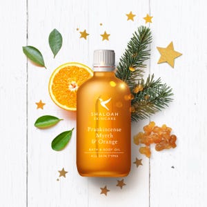 Peut inclure: Une bouteille ambrée d'huile pour le bain et le corps Frankincense, Myrrhe & Orange de Shaloah Skincare. L'image comprend une tranche d'orange, des feuilles vertes, des étoiles dorées, une branche de conifère et des morceaux de résine. Pour tous types de peau.